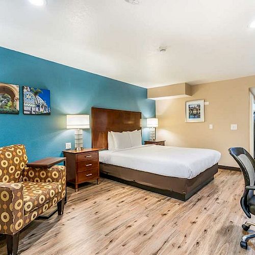 A well-lit hotel room featuring a bed, a patterned armchair, desk, chair, and wall art, with wooden flooring and modern furnishings.