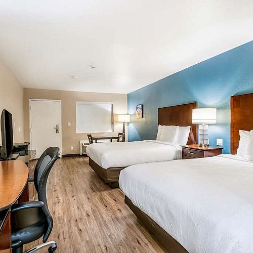 The image shows a modern hotel room with two beds, a desk, a chair, and a TV. The room has light walls, wooden flooring, and a blue accent wall.