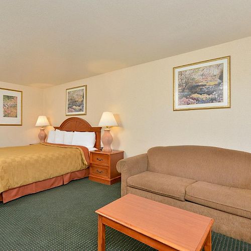 The image shows a hotel room with a large bed, two bedside tables with lamps, a sofa, a small coffee table, and framed artwork on the walls.