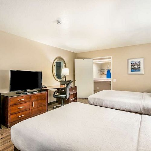 A hotel room with two beds, a TV on a dresser, a desk with a chair, and a small fridge. There's a lamp and wall art in the background.