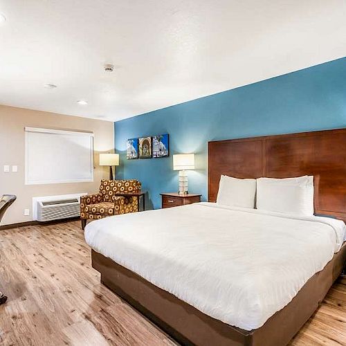 A modern hotel room with a large bed, bedside tables, lamps, a TV, a desk with a chair, an armchair, and wood flooring, featuring a blue accent wall.