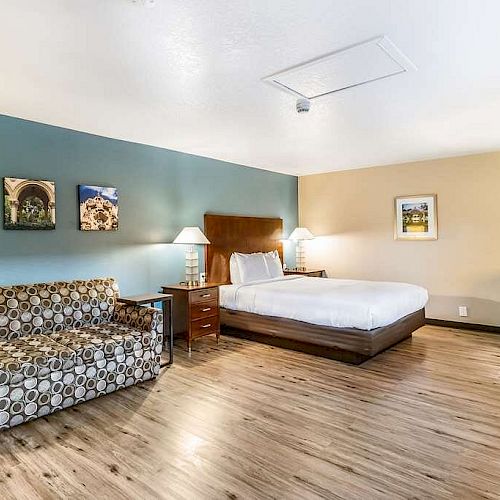 A modern hotel room featuring a bed, a patterned sofa, a work desk with a chair, and wall art. The floor is wooden, and the lighting is subtle.