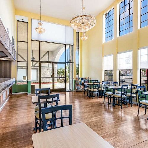 The image shows a bright, spacious dining area with large windows, chandeliers, and several tables and chairs. There's a counter on the left side.