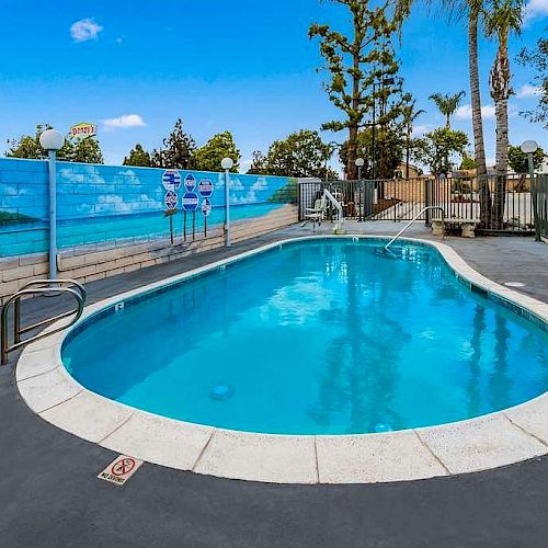 An outdoor swimming pool is surrounded by a gated fence, with a mural and safety equipment on the wall. Trees and a clear blue sky are visible.