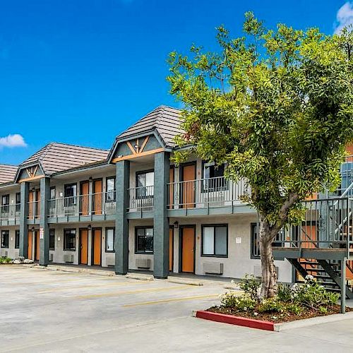 This image shows a modern, two-story apartment complex with outdoor staircases, parking area, and a tree in the foreground under a clear blue sky.