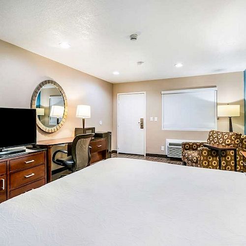 A hotel room with a bed, flat-screen TV on a dresser, desk with chair, armchair with lamp, and wall-mounted mirror. The room has a calming, clean design.