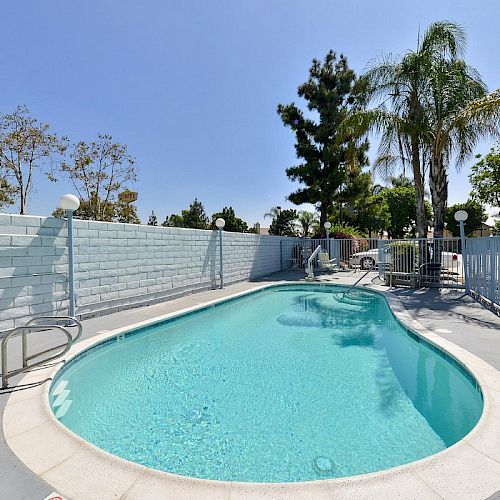 This image shows an outdoor swimming pool with a curved shape, surrounded by a concrete patio and a fence, with trees and a clear sky in the background.