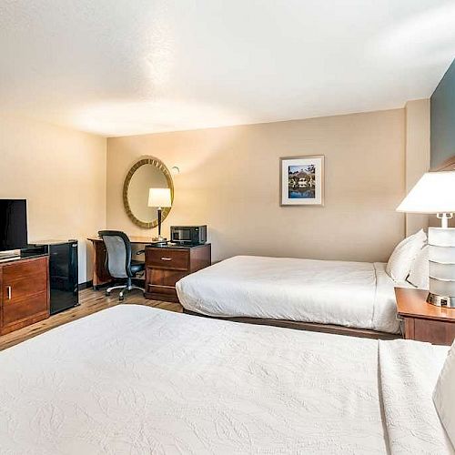 The image shows a hotel room with two beds, a dresser with a TV, a desk with a chair, a mirror, and lamps, all in a neatly arranged setup.