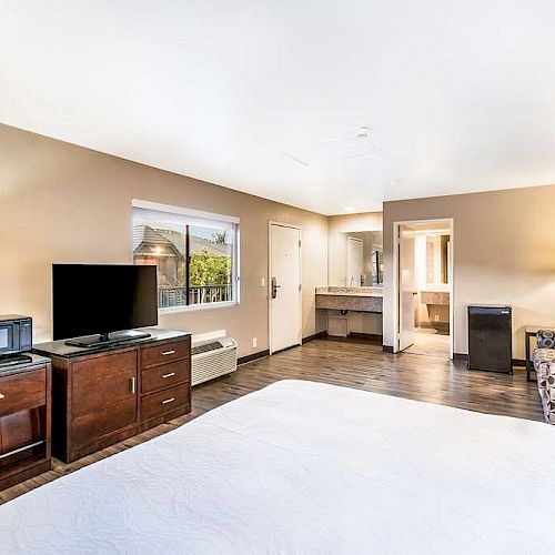 This image shows a tidy hotel room with a bed, TV, microwave, and fridge. There's also a couch, a lamp, and a clean, well-lit bathroom door.