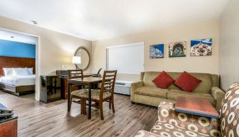 The image shows a well-furnished hotel room with a seating area, dining table, and a bed in a separate space. There are three artworks on the wall.